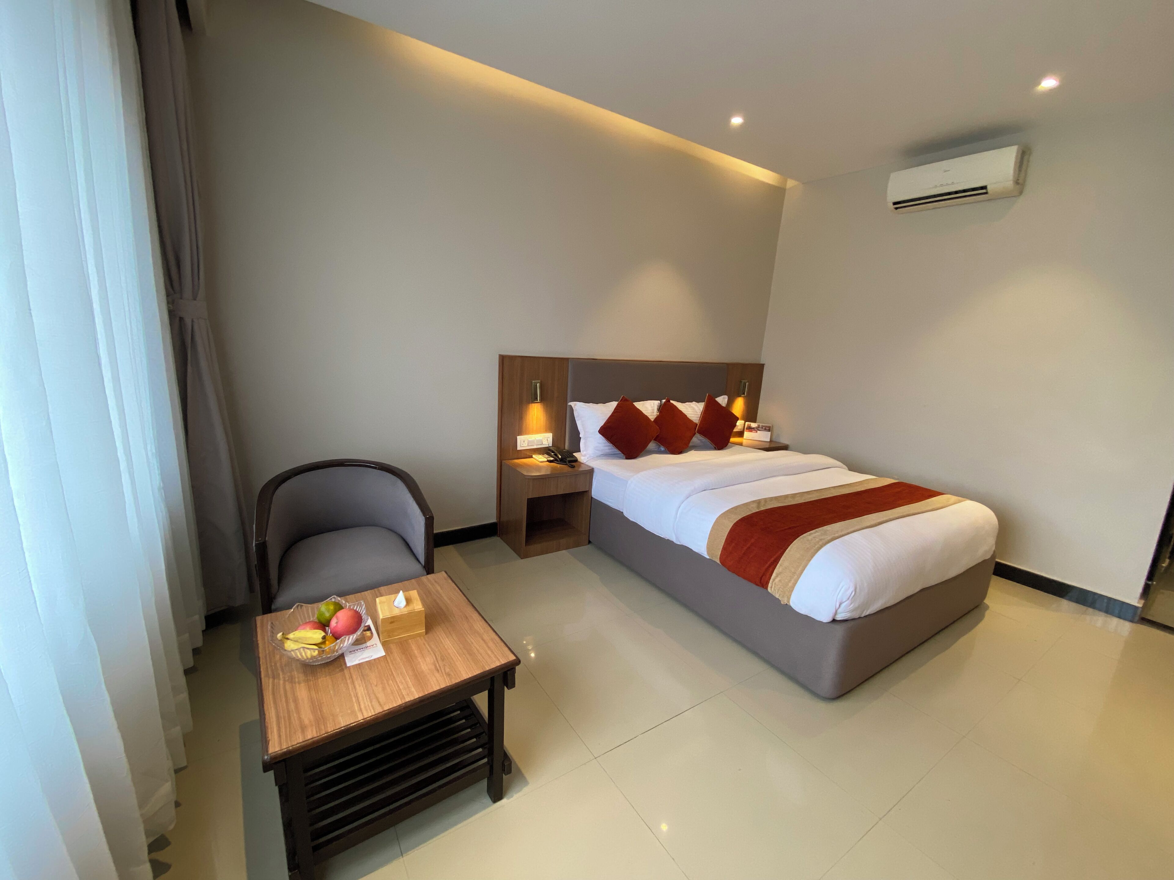 A hotel room with a bed, bedside table, chair, and a small table with fruits and a tissue box.