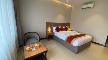Habitación de hotel con cama, mesita de noche, silla y una pequeña mesa con frutas y una caja de pañuelos.