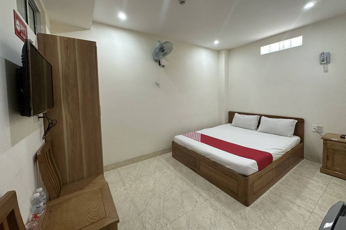 A bedroom with a wooden bed, a TV mounted on the wall, a ceiling fan, and a small window.
