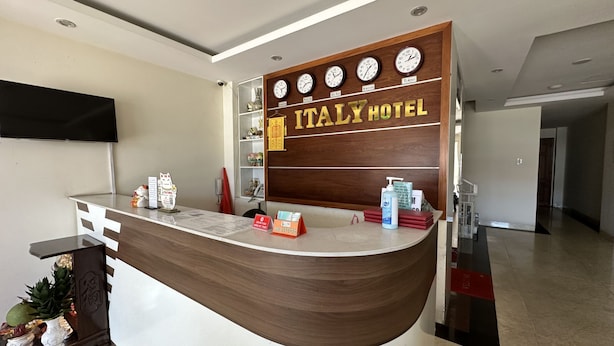 A reception desk with clocks showing different time zones, a sign for Italy Hotel, and a counter with various items. - Italy Hotel (Nam Nha Trang)