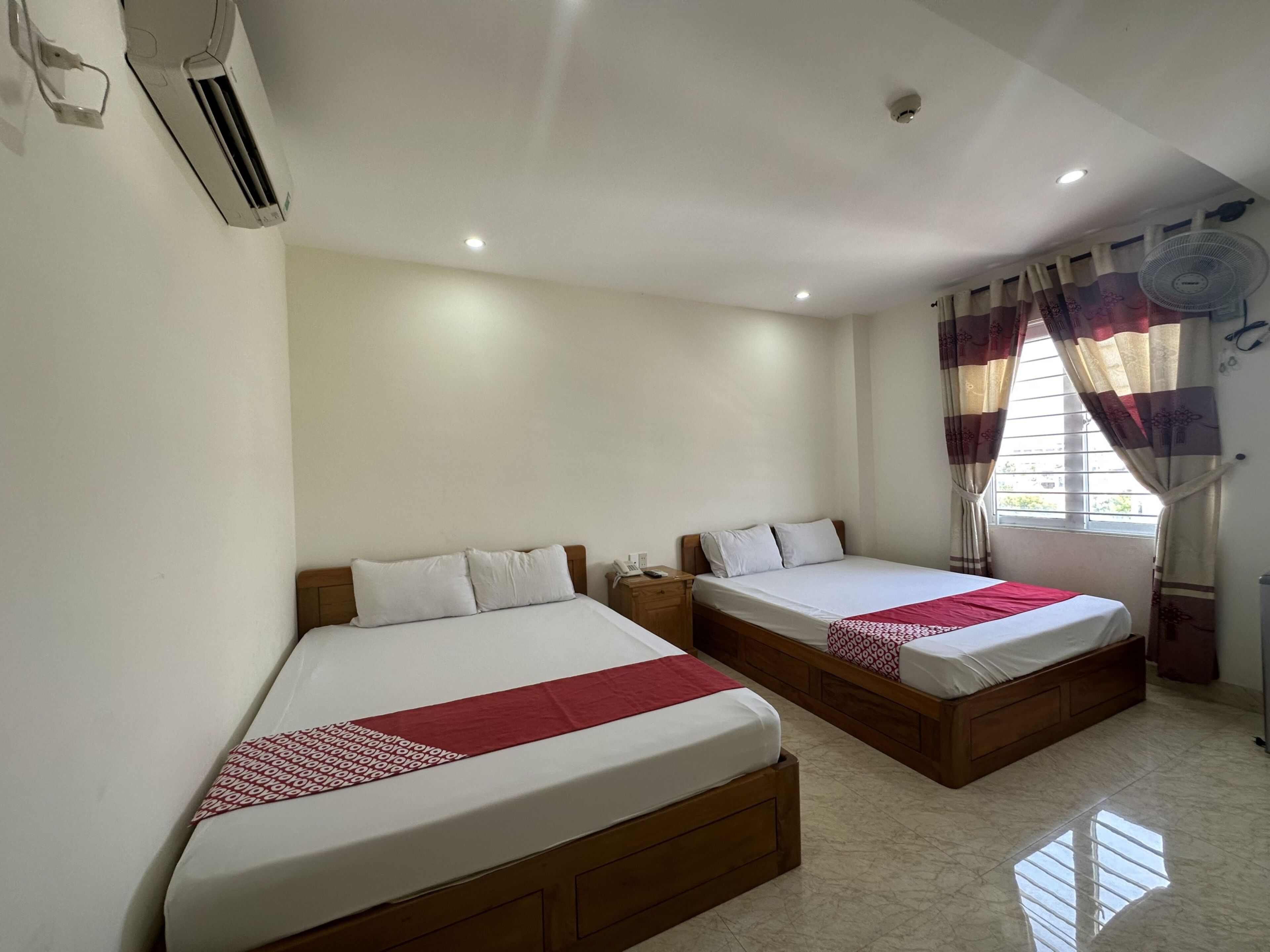 A hotel room with two beds, a ceiling fan, an air conditioning unit, and a window with curtains.