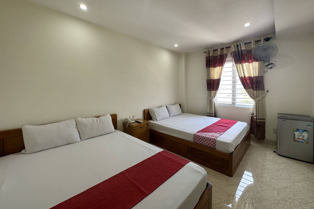 A hotel room with two beds, a small table, a fan, and a refrigerator.