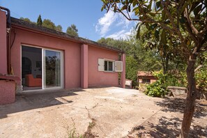 Un bâtiment rose avec une porte et une fenêtre en verre, une terrasse en pierre et de la verdure tout autour.