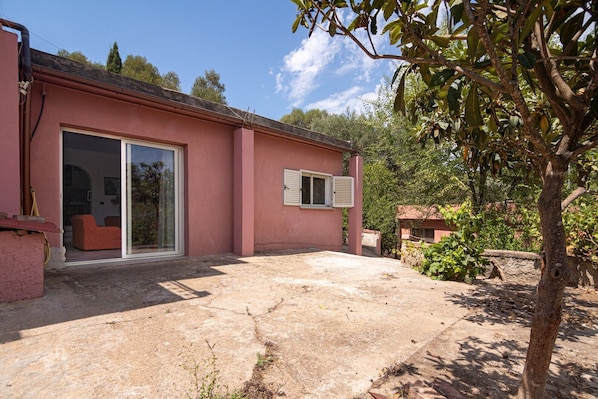 A pink building with a glass door and window, a stone patio, and surrounding greenery.
