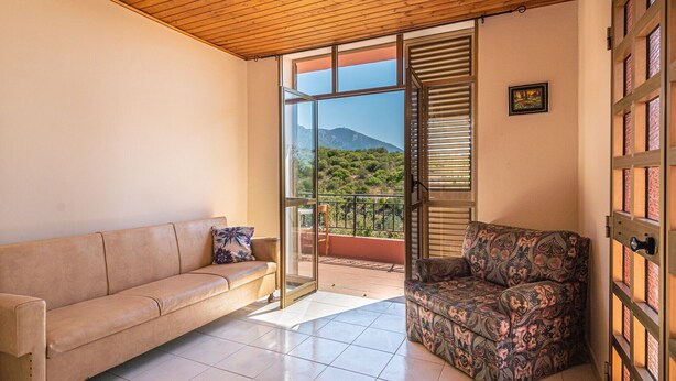 A room with a beige sofa, a patterned armchair, a glass door leading to a balcony with a mountain view, and a wooden ceiling. - Holiday Home 'Casa Teresa' with Mountain View, Balcony, and Air Conditioning (Iglesias)