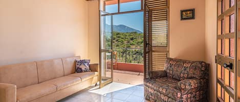 A room with a beige sofa, a patterned armchair, a glass door leading to a balcony with a mountain view, and a wooden ceiling.
