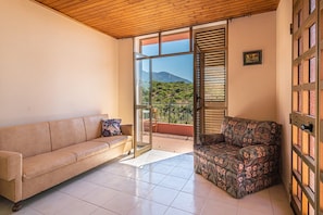 Une pièce avec un canapé beige, un fauteuil à motifs, une porte en verre donnant sur un balcon avec vue sur la montagne et un plafond en bois.