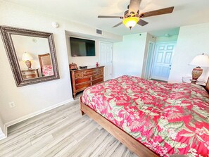 Ein Schlafzimmer mit Bett, Kommode, Spiegel, Deckenventilator und einer Tür zu einem weiteren Raum.