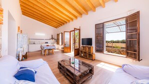 A spacious living area with a wooden ceiling, a glass coffee table, and a view of a garden through open windows.