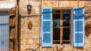 A rustic stone building with a wooden door and blue shutters, a small window with a lantern, and a potted plant.