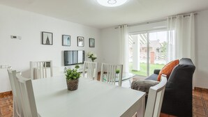 Una sala de estar moderna con una mesa de comedor, sillas blancas, un sofá, un televisor de pantalla plana y una planta en maceta.