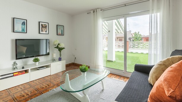 Una sala de estar con televisor, una mesa de centro de vidrio, un sofá y una puerta corrediza de vidrio hacia un jardín.