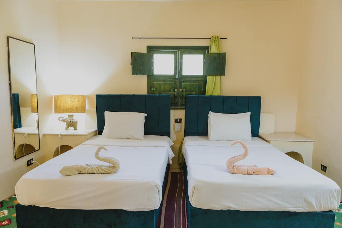 A hotel room with two single beds, a large mirror, a window with curtains, and a bedside table with a lamp.