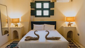 Habitación de hotel con cama, dos lámparas de noche, espejo, ventana con contraventanas y equipo de aire acondicionado.