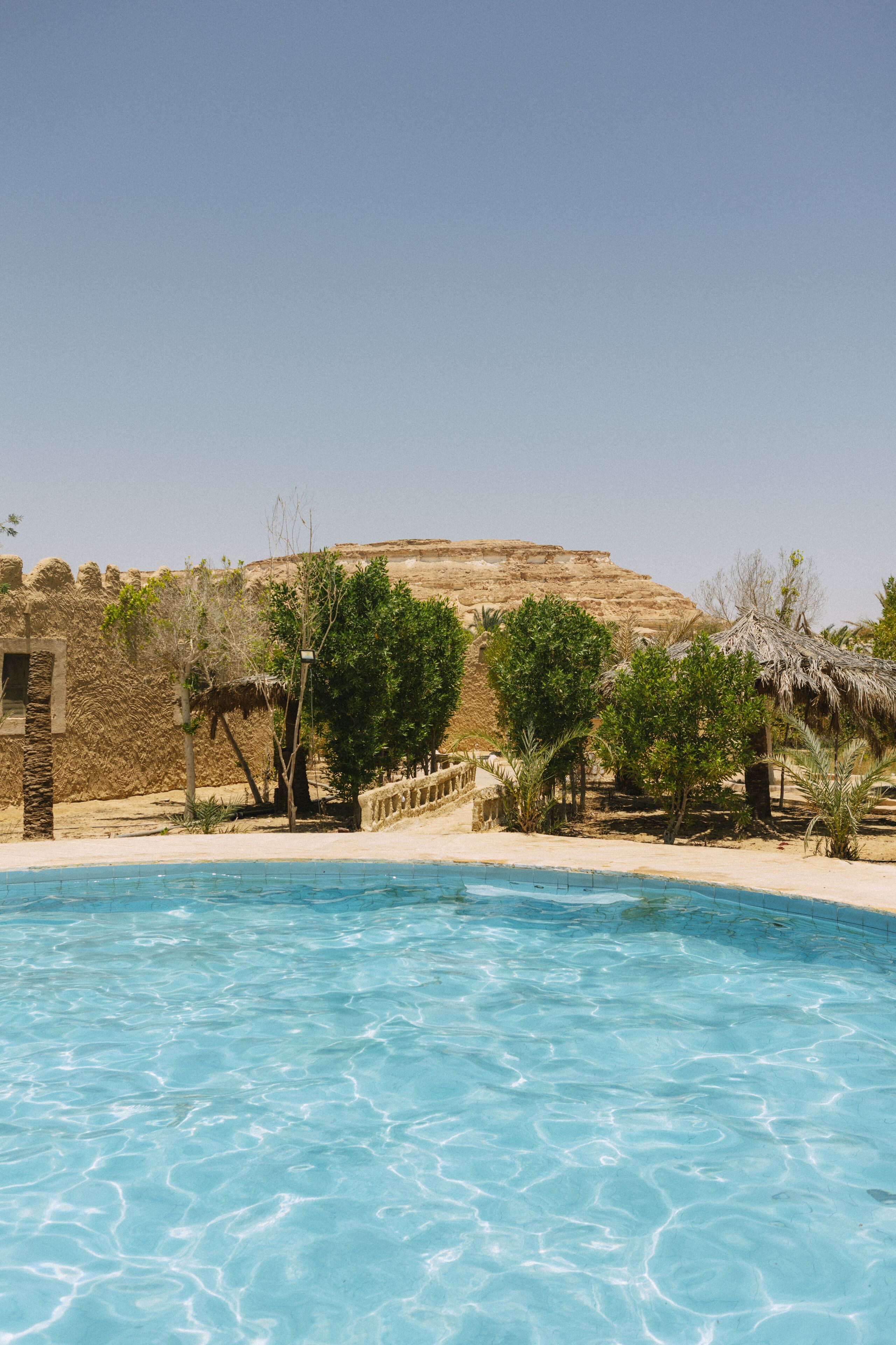 Uma piscina com água azul cristalina, rodeada por uma área de areia com árvores e uma parede de pedra.