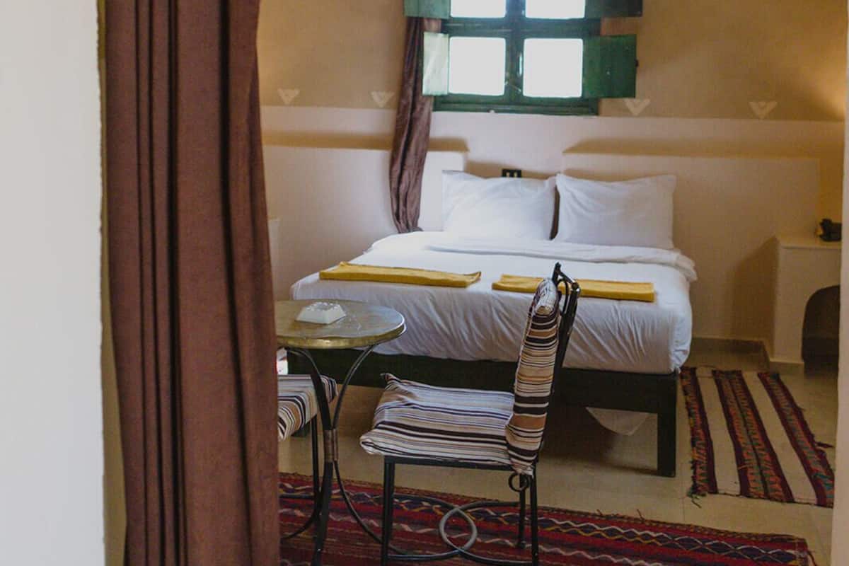 A bedroom with a bed, a chair, a small table, a ceiling fan, and a window with shutters.