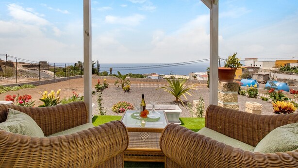 A covered outdoor seating area with wicker furniture, a table with a bottle and glass, and a view of the sea. - Vacation Home 'Finca Mariposa' with Sea View, Garden & Wi-Fi (Adeje)