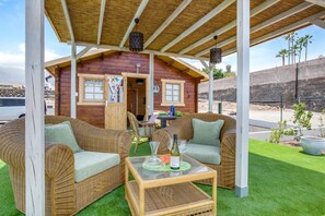 Une cabane en bois dotée d’un patio couvert, de meubles en osier, d’une table avec une bouteille et un verre, et d’un petit espace aménagé en jardin.