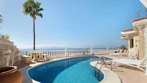 A swimming pool with a view of the sea, white lounge chairs, and a palm tree.
