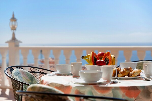 Une table dressée avec des aliments pour le petit-déjeuner, notamment des fruits et des viennoiseries, sur un balcon doté d’une balustrade, avec un lampadaire en arrière-plan.