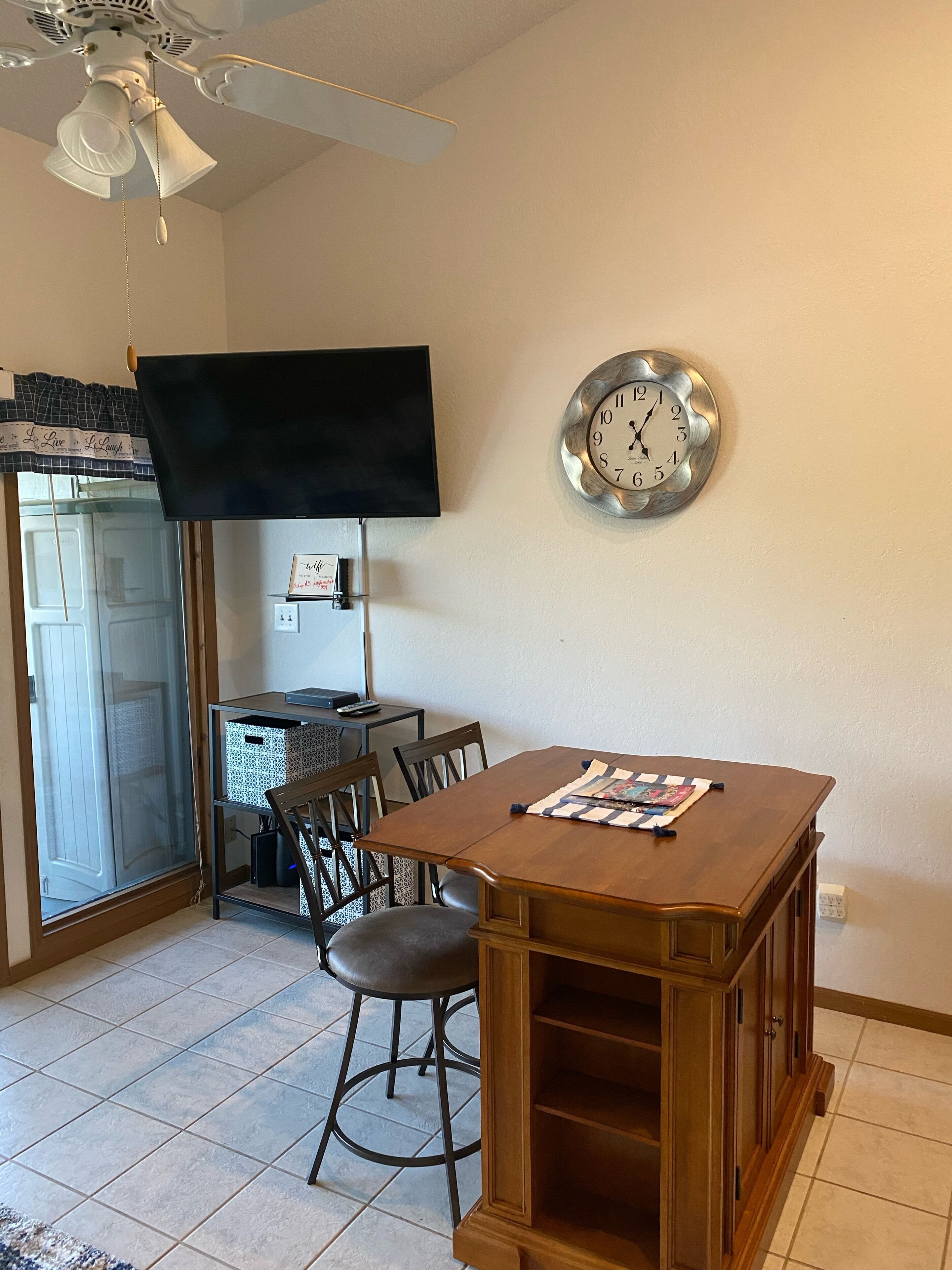 Comedor con mesa de madera, sillas, un reloj, televisor y ventilador de techo.