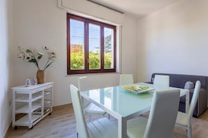 A dining area with a glass table, white chairs, a white cart with shelves, a window with a view of greenery, and a grey sofa.