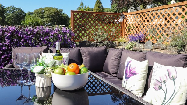 A glass table with a bowl of fruit and wine glasses, a white vase with flowers, and a cushioned outdoor seating area with purple cushions.