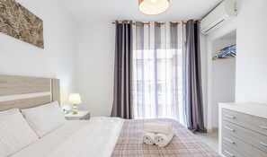 A hotel room with a bed, a nightstand with a lamp, a dresser, and a window with curtains.