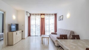 Ein kompakter Wohnraum mit Sofa, einem kleinen Tisch, einem Fernseher auf einem Schrank und einem Balkon mit Vorhängen.