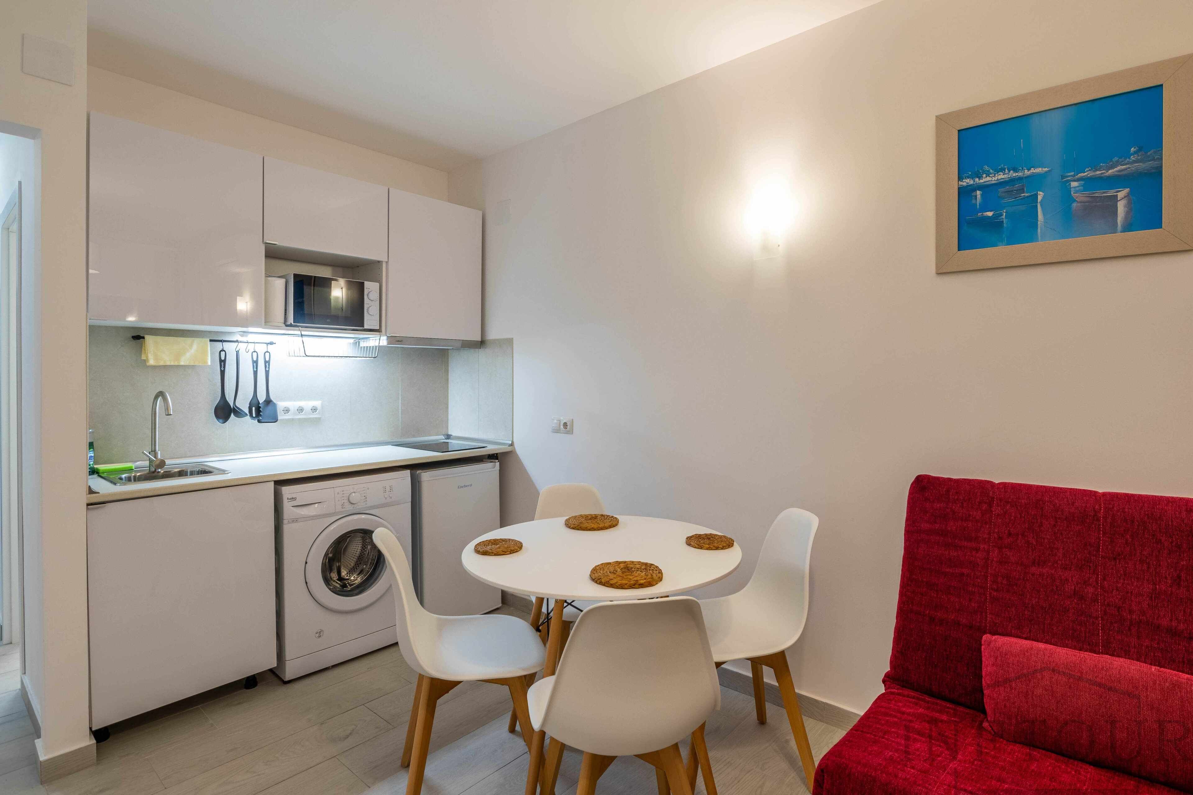 A compact kitchen with white cabinets, a washing machine, and a dining area with a red sofa and white chairs.