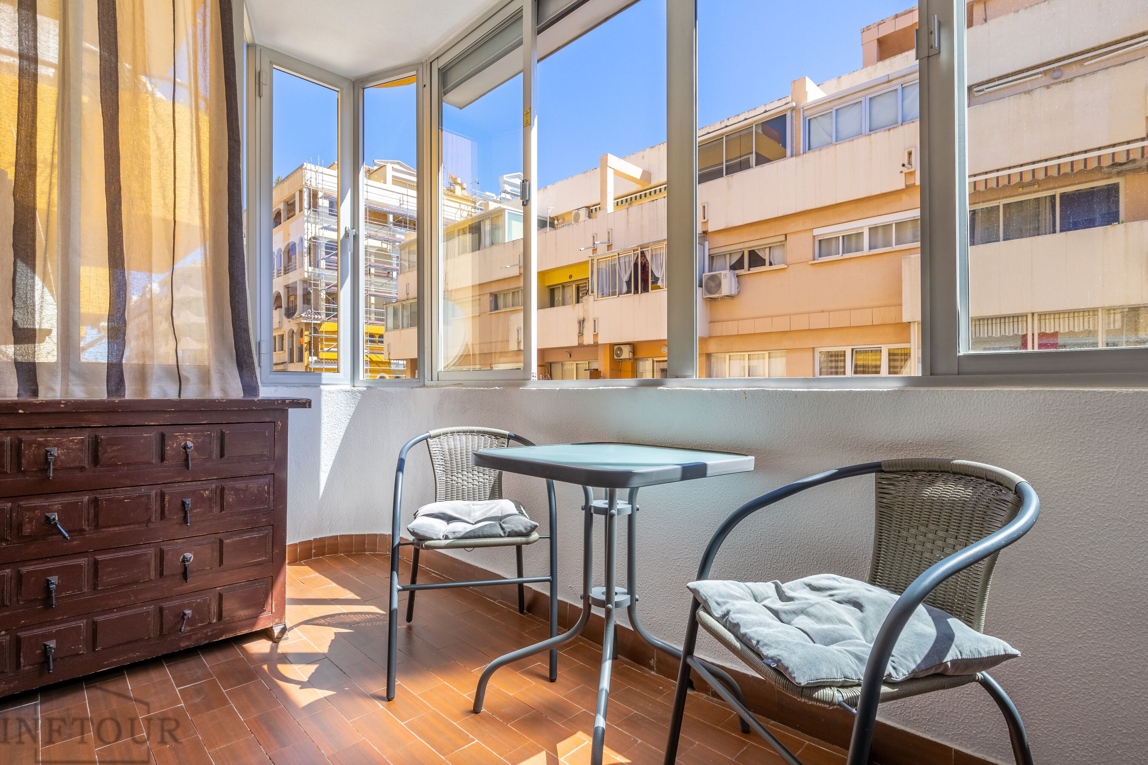 Ein Balkon mit einem Holzkasten, zwei Korbstühlen, einem kleinen Tisch und Blick auf mehrstöckige Gebäude.