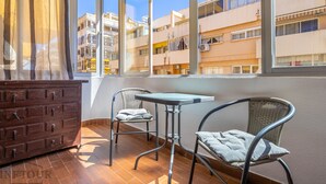 Ein Balkon mit einem Holzkasten, zwei Korbstühlen, einem kleinen Tisch und Blick auf mehrstöckige Gebäude.