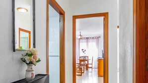 A hallway leading to a dining area with a table and chairs, a vase with flowers on a console table, and a refrigerator.