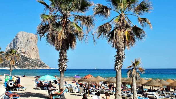 A beach with palm trees, beach chairs, umbrellas, and a rocky cliff in the background. - Inftour Stefano Apartamento (Calp)