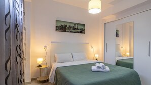 A hotel room with a bed, a nightstand, a lamp, a mirror, and a cityscape painting on the wall. - Inftour Stefano Apartamento (Calp)