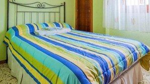 Een enkel bed met een gestreept dekbedovertrek, een plafondventilator, een houten deur en een raam met dunne gordijnen.