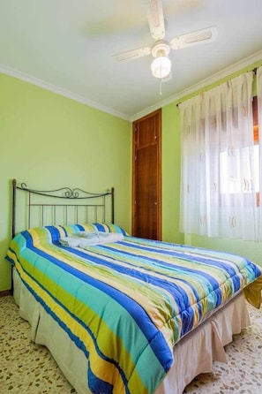 A single bed with a striped bedspread, a ceiling fan, a wooden door, and a window with sheer curtains.