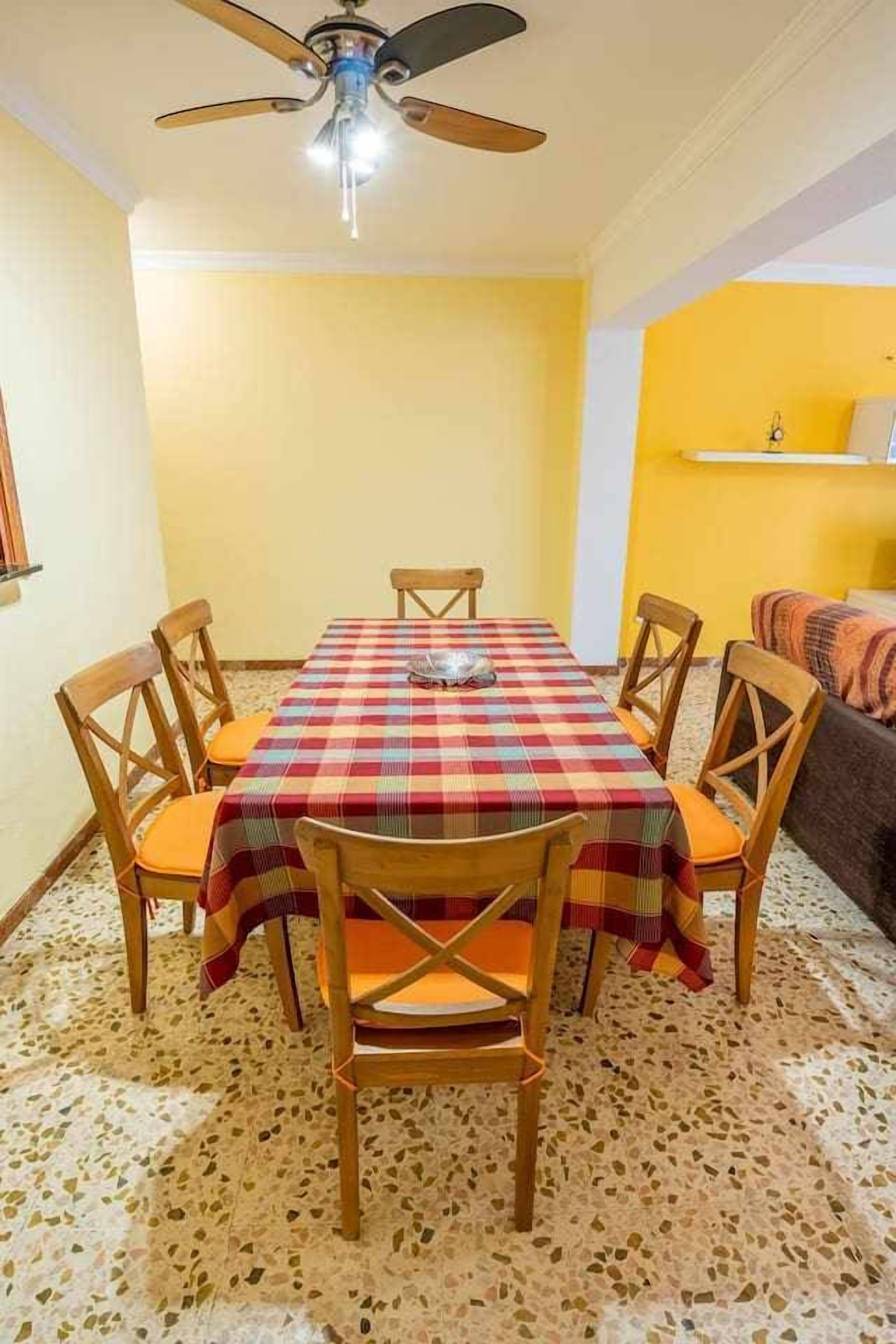 A dining area with a table covered by a checkered tablecloth, surrounded by wooden chairs with orange cushions. A ceiling fan is visible above.