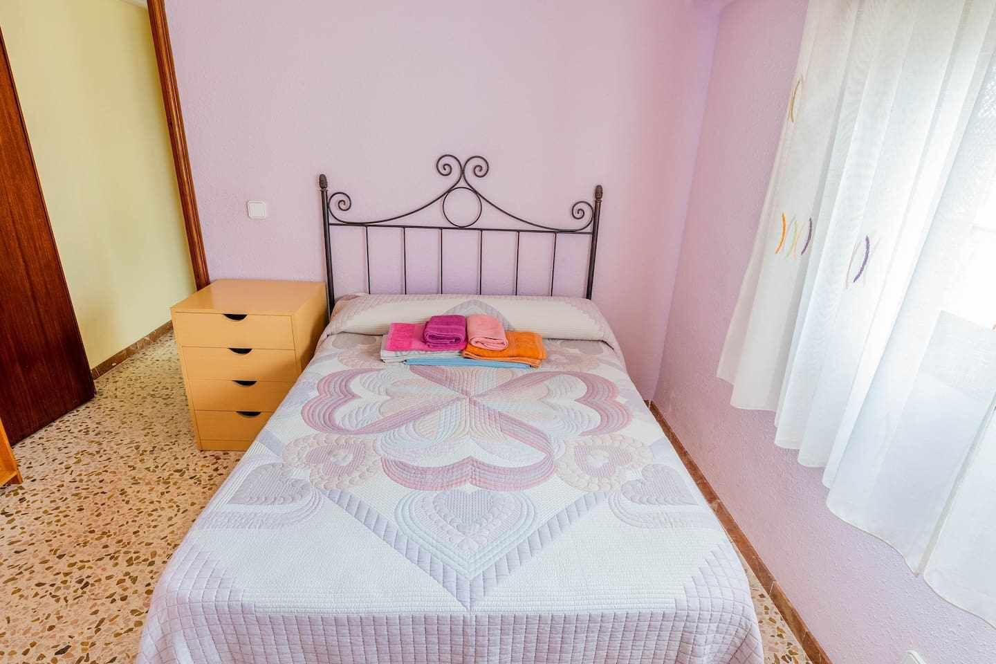 Ein Einzelbett mit Metallgestell, einer gemusterten Bettdecke und einem Satz gefalteter Handtücher auf dem Bett.