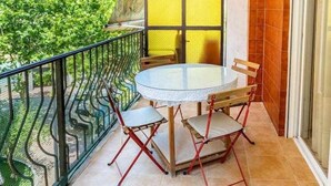 Een balkon met een ronde glazen tafel en vier stoelen, een geel afscheiding en uitzicht op gebouwen en groen.