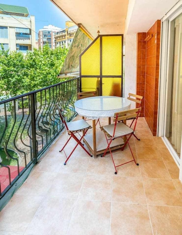 Un balcón con una mesa redonda de vidrio y cuatro sillas, una separador amarillo y vistas a edificios y zonas verdes.