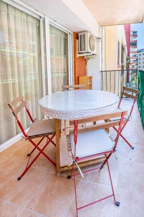 Una mesa redonda de vidrio con cuatro sillas de madera en un balcón con suelo de baldosas y vistas a edificios.