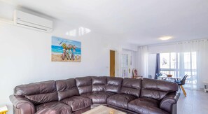A living room with a brown leather sectional sofa, a painting of a beach scene, and a dining area with a table and chairs.
