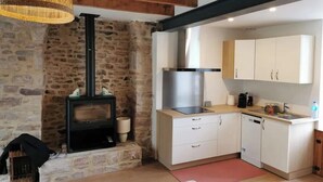 A cozy room with a stone wall, a wood-burning stove, a modern kitchen with white cabinets, and a hanging light fixture. - Charming townhouse, 10 minutes walk from the beach! (Erquy)