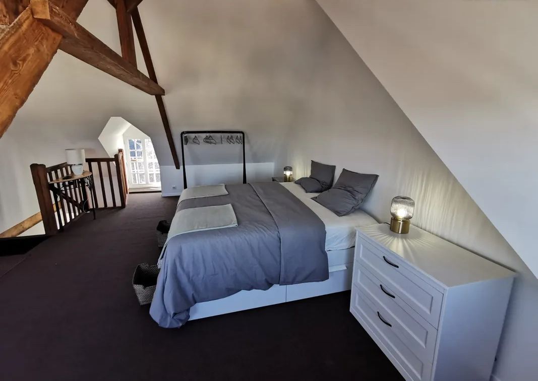 Een slaapkamer met een schuine plafond, een bed met grijze beddengoed, een nachtkastje met een lamp, en een witte dressoir.