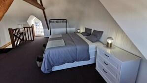 Une chambre avec un plafond en pente, un lit avec une literie grise, une table de chevet avec une lampe et une commode blanche.