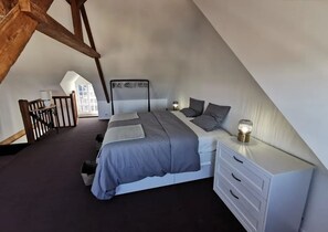 A bedroom with a sloped ceiling, a bed with grey bedding, a bedside table with a lamp, and a white dresser. - Charming townhouse, just a 10-minute walk from the beach! (Erquy)