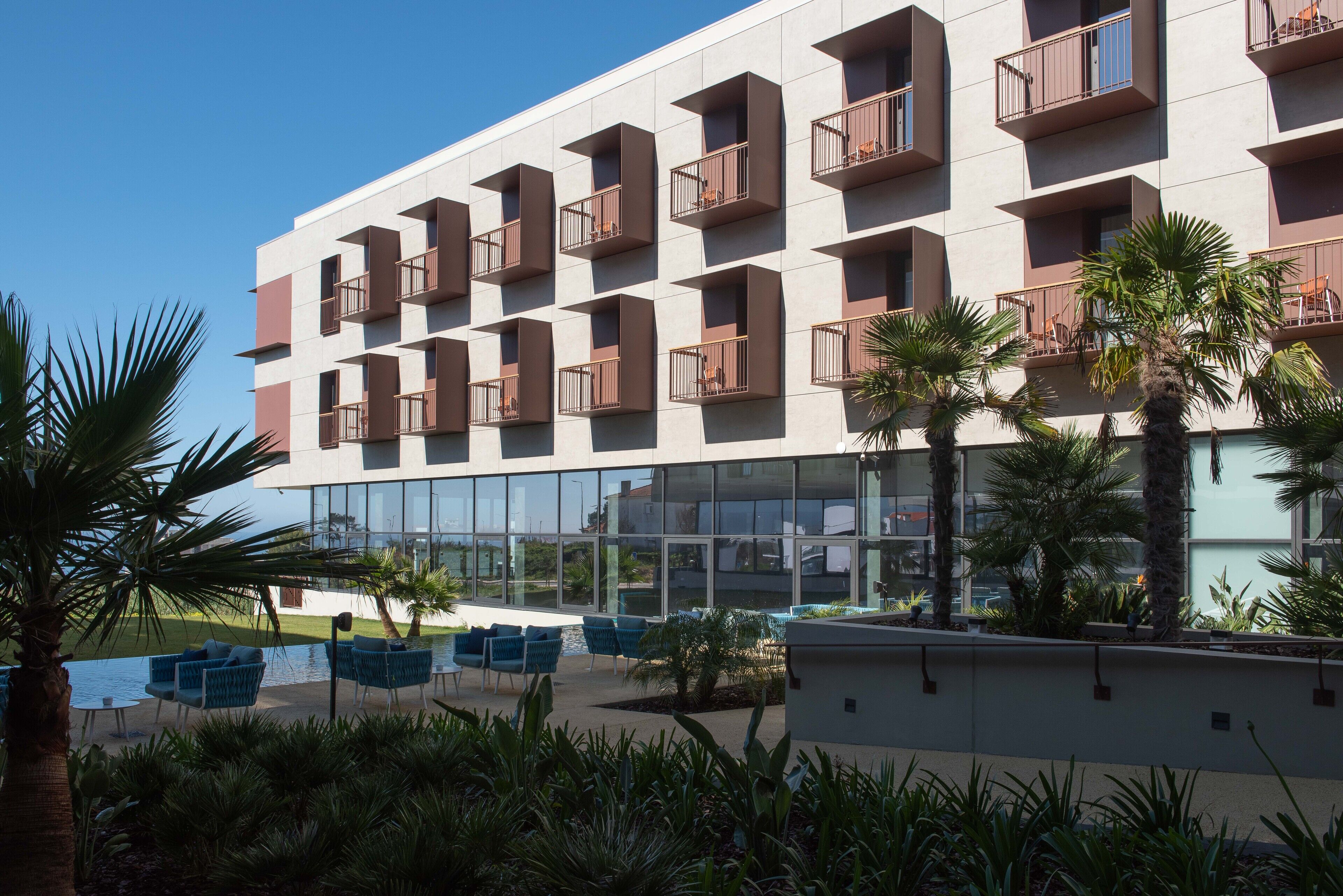 Un hotel modern amb múltiples balcons, mobiliari exterior i palmeres.