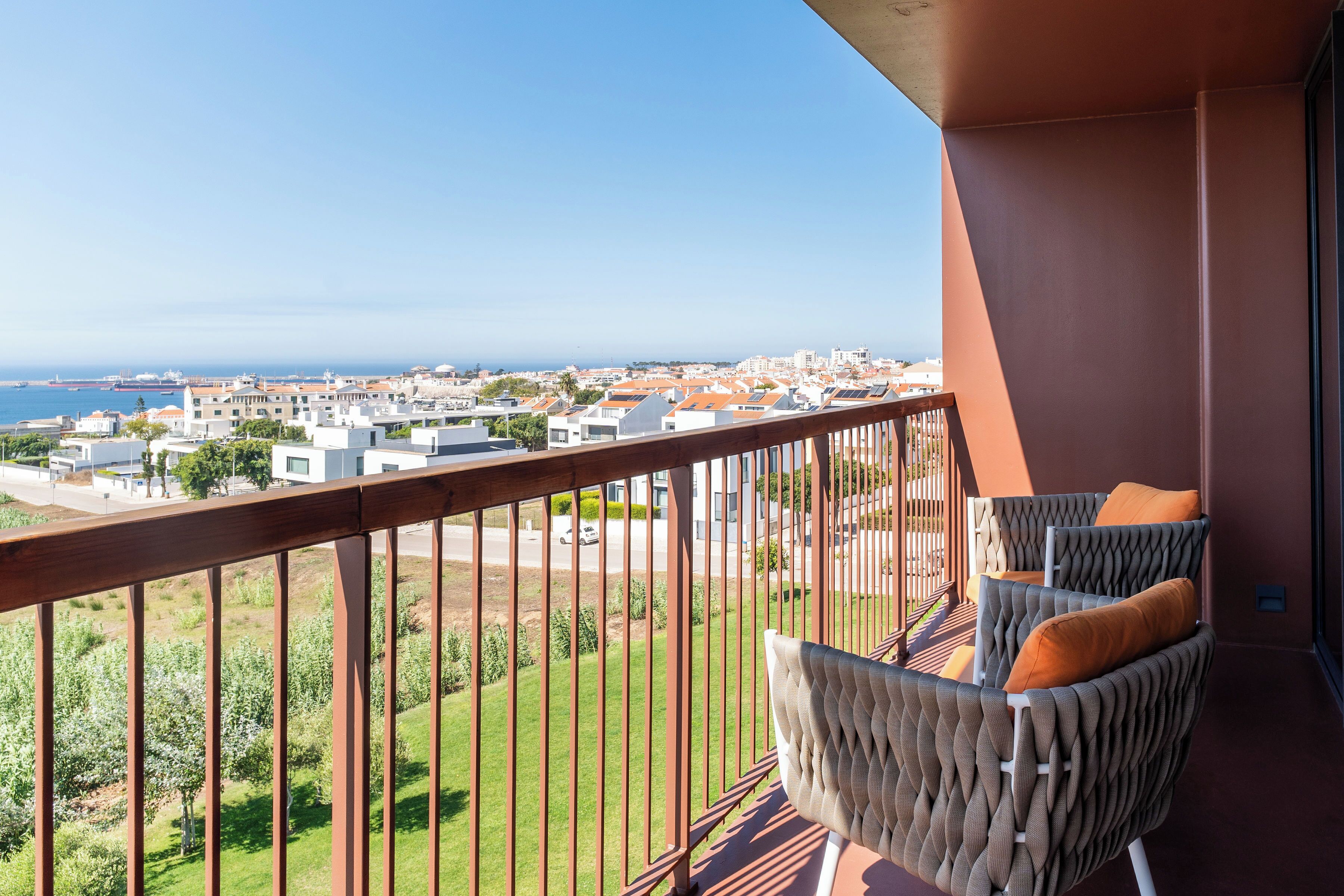 A balcony with a view of a coastal town, featuring a seating area with cushions.