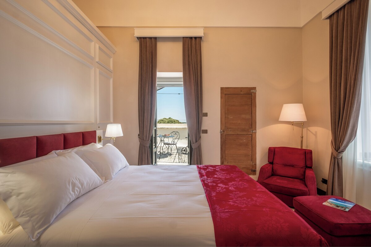 A hotel room with a large bed, a red armchair, a small table with a lamp, and a view of the outdoors through a door.